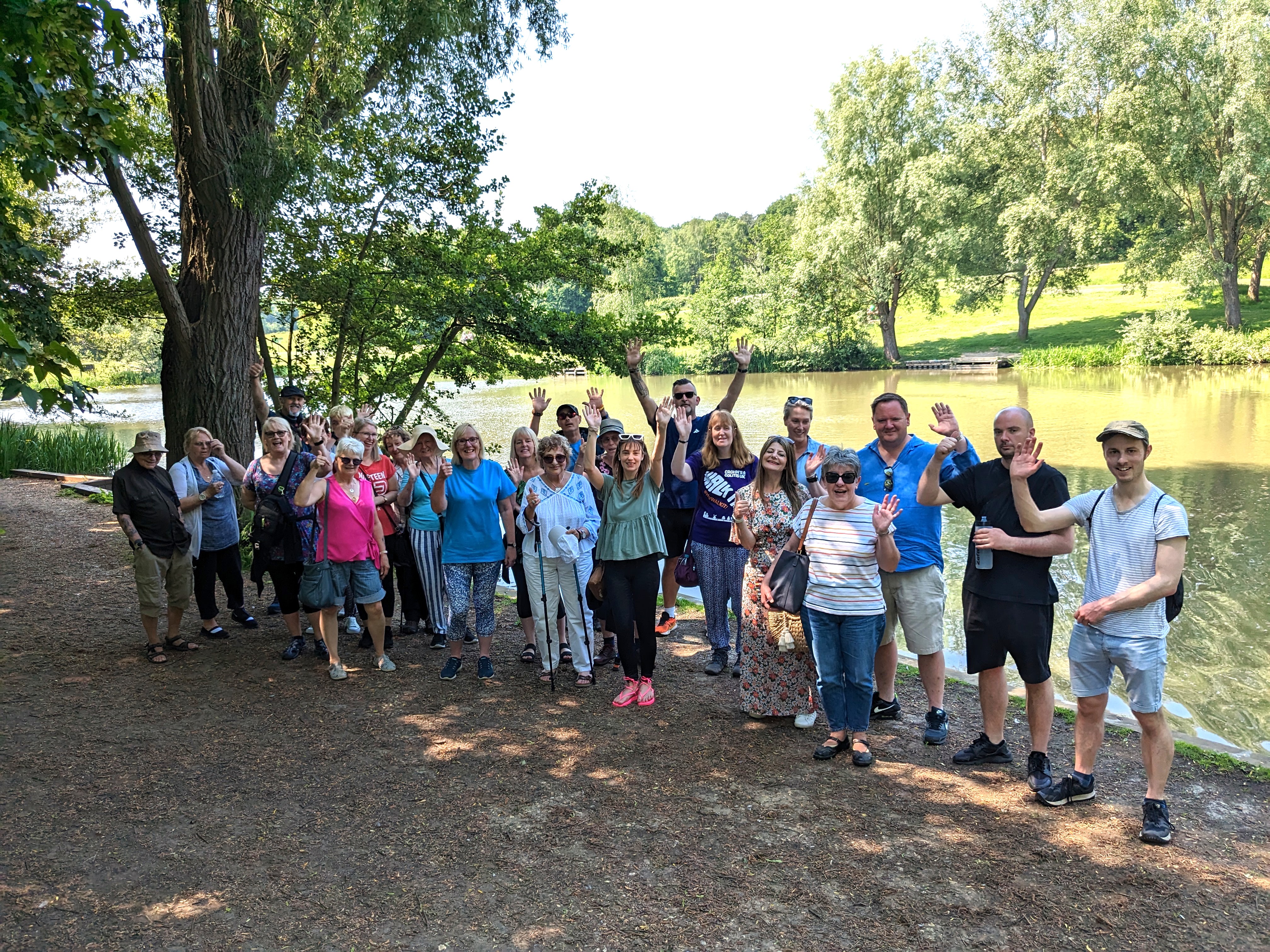 A groups of people stand in a parkland area, on a sunny day, smiling and waving at the camera