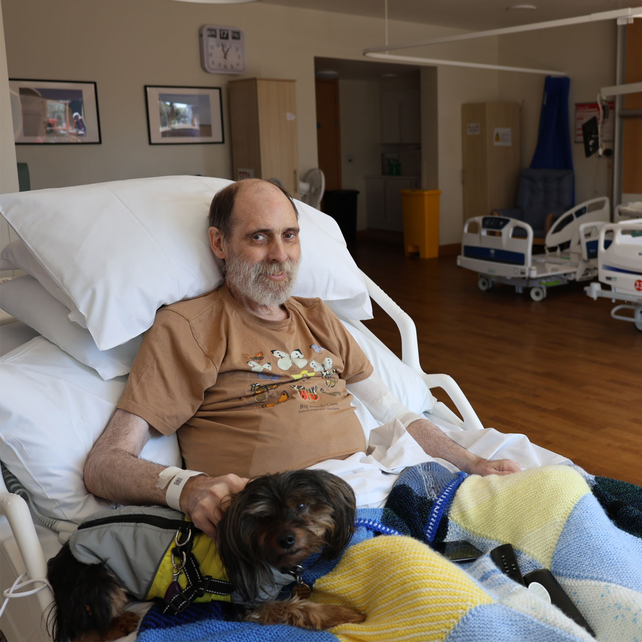 Bradley sits in his St Helena Hospice bed smiling at the camera with his little dog on the bed next to him.